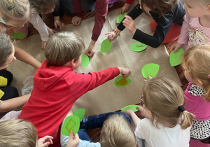 Dzieci na dywanie przyklejają zielone listki do narysowanego na papierze drzewa