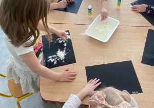 Dzieci przy stoliku rozsypują mąkę na czarne kartony pomalowane klejem