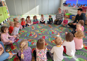 Dzieci z nauczycielką i autorem książki siedzą w kole na dywanie, Słuchają opowiadania czytanego przez autora