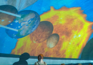 Dzieci z nauczycielką siedzą pod kopułą mobilnego planetarium i oglądają pokaz dotyczący układu słonecznego