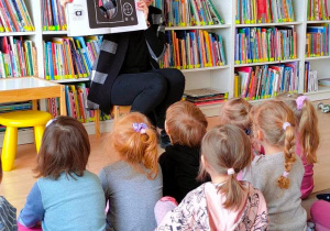 Dzieci przyglądają się ilustracji aparatu fotograficznego w "Książce z dziurą". Książkę trzyma Pani bibliotekarka