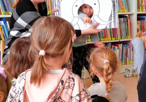 Dzieci obserwują dziewczynkę, która swoją ręką naśladuje ruchy trąby słonia. Ręka dziewczynki przełożona jest przez dziurę w ilustracji książki