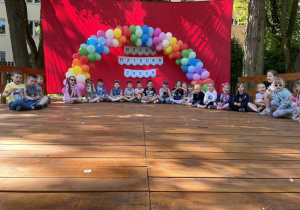 dzieci siedzą na scenie pod łukiem z balonów