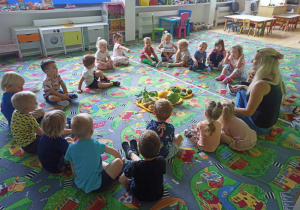 Sala przedszkolna. Dzieci siedzą na dywanie w kole. Na środku koła leżą warzywa. Dzieci rozmawiają z nauczycielką o warzywach.