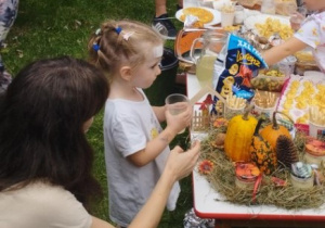 Ogród przedszkolny. W tle napis "Wiewiórki" .Dzieci i rodzice na pikniku rodzinnym zgromadzeni przy stole, na którym znajdują się potrawy przygotowane przez rodziców.