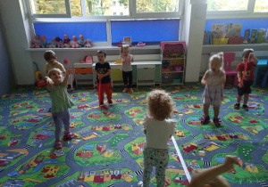 Sala przedszkolna. Dzieci na dywanie biorą udział w zabawach muzyczno-ruchowych z wstążkami zrobionymi z czerwonej bibuły.