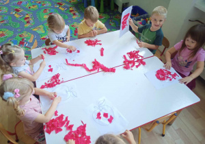 Sala przedszkolna. Dzieci siedzą przy stole, wykonują pracę plastyczną. Wyklejają kontur jabłka, kawałkami czerwonej bibuły.