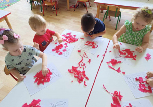 Sala przedszkolna. Dzieci siedzą przy stole, wykonują pracę plastyczną. Wyklejają kontur jabłka, kawałkami czerwonej bibuły.