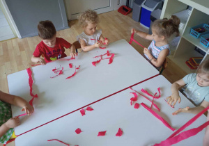 Sala przedszkolna. Dzieci siedzą przy stole, drą paski czerwonej bibuły na mniejsze kawałki.