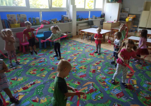 Sala przedszkolna. Dzieci na dywanie biorą udział w zabawach muzyczno-ruchowych z wstążkami zrobionymi z czerwonej bibuły.