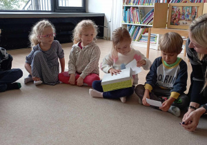 Dzieci i pani bibliotekarka obserwują dziewczynkę losującą z pudełka karteczkę z dobrymi słowami