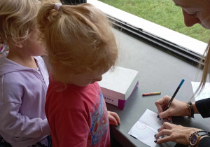 Dziewczynka przygląda się pani bibliotekarce piszącej na karteczce dobre słowa podyktowane przez dziewczynkę