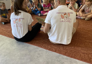 Dzieci na dywanie przed nimi Pan i Pani z wydawnictwa Molly czytają dzieciom książki