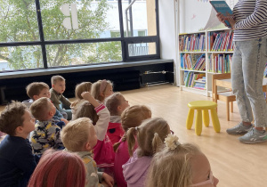 Dzieci w sali bibliotecznej słuchają czytanej przez bibliotekarkę książki.