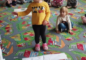 Sala przedszkolna. Dzieci siedzą na dywanie. Dziewczynka stoi przed przejściem dla pieszych, oznaczonym na dywanie przez białe pasy wycięte z papieru i wskazuje ręką jak należy rozejrzeć się przed przejściem na drugą stronę jezdni.