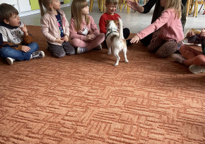 Dzieci na dywanie słuchają Pani, pies podbiega do chłopca