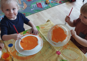 Sala przedszkolna. Dzieci siedzą przy stoliku, malują papierowe talerze pomarańczową farbą, używając pędzli.