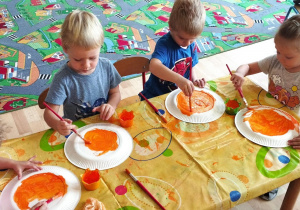Sala przedszkolna. Dzieci siedzą przy stoliku, malują papierowe talerze pomarańczową farbą, używając pędzli.