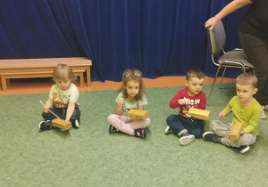Sala gimnastyczna. Dzieci siedzą na podłodze, grają na różnych instrumentach.