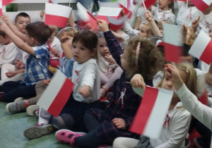 Sala gimnastyczna. Dzieci biorą udział w uroczystości z okazji Święta Niepodległości. Dzieci siedzą na podłodze, trzymają flagi Polski.
