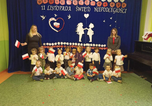 Sala gimnastyczna. Dzieci z panią dyrektor, wychowawczynią pozują do zdjęcia na tle dekoracji przygotowanej z okazji Święta Niepodległości.