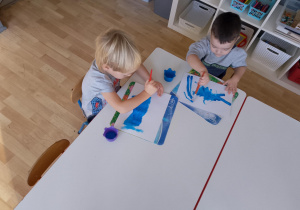 Sala przedszkolna. Dwóch chłopców siedzi przy stoliku, malują karton niebieską farbą z użyciem pędzla.