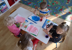 Sala przedszkolna. Dzieci siedzą przy stoliku, malują karton farbami z użyciem pędzla.
