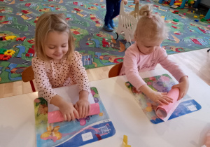 Sala przedszkolna. Dwie dziewczynki siedzą przy stoliku i zwijają pomalowane kartony.