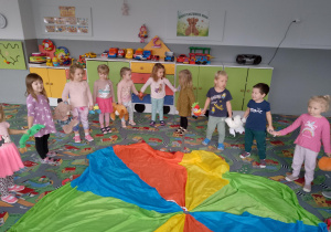 Sala przedszkolna. Dzieci ustawione w kole wiązanym, dookoła chusty animacyjnej trzymają pluszaki.