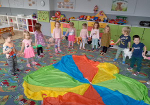 Sala przedszkolna. Sala przedszkolna. Dzieci ustawione w kole wiązanym, dookoła chusty animacyjnej trzymają pluszaki.