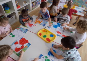 Sala przedszkolna. Dzieci siedzą przy stolikach, malują farbami sylwetki misia.