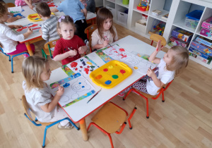 Sala przedszkolna. Sala przedszkolna. Dzieci siedzą przy stolikach, malują farbami sylwetki misia.