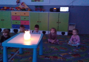 Sala przedszkolna. ala przedszkolna. Dzieci siedzą na dywanie dookoła stołu, na którym stoi świeca.