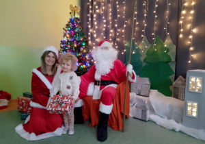 Sala gimnastyczna. Dziewczynka trzyma prezent, obok dziewczynki siedzi św. Mikołaj, a z drugiej strony Śnieżynka. W tle dekoracja świąteczna.