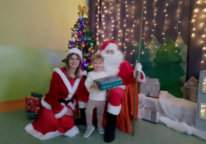 Sala gimnastyczna. Chłopiec trzyma prezent, obok chłopca siedzi św. Mikołaj, a z drugiej strony Śnieżynka. W tle dekoracja świąteczna.