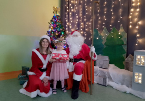 Sala gimnastyczna. Dziewczynka trzyma prezent, obok dziewczynki siedzi św. Mikołaj, a z drugiej strony Śnieżynka. W tle dekoracja świąteczna.