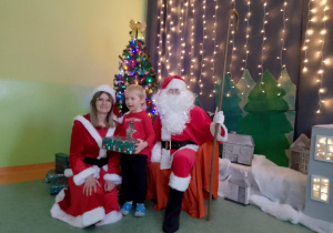 Sala gimnastyczna. Chłopiec trzyma prezent, obok chłopca siedzi św. Mikołaj, a z drugiej strony Śnieżynka. W tle dekoracja świąteczna.