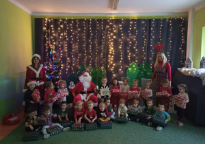 Sala gimnastyczna. Dzieci wraz ze św. Mikołajem, Śnieżynką i wychowawczynią pozują do zdjęcia na tle dekoracji świątecznej.