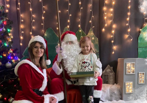Dziewczynka z prezentem w ręku pozująca do zdjęcia ze Świętym Mikołajem i Panią Mikołajową