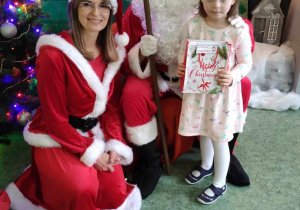 Dziewczynka, Mikołaj i Mikołajka pozują do zdjęcia na tle świątecznej dekoracji