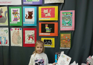 Dziewczynka pozuje do zdjęcia na tle prac plastycznych przedstawiających koty