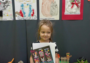 Dziewczynka pozuje do zdjęcia na tle prac plastycznych przedstawiających koty