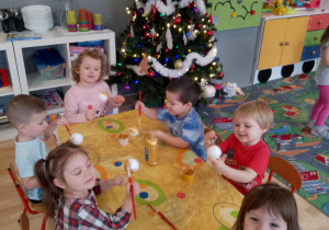 Sala przedszkolna. Dzieci siedzą przy stoliku i malują farbami świąteczne ozdoby.