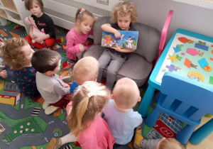 Sala przedszkolna. Dziewczynka siedzi na kanapie i pokazuje, dzieciom siedzącym przed nią, książkę..