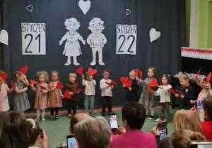Sala gimnastyczna. Dzieci wykonują taniec z czerwonymi serduszkami, wyciętymi z papieru. Za nimi dekoracja z okazji Dnia Babci i Dziadka.