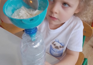 Sala przedszkolna. Dziewczynka za pomocą łyżki i lejka, wsypuje mąkę do plastikowej butelki.