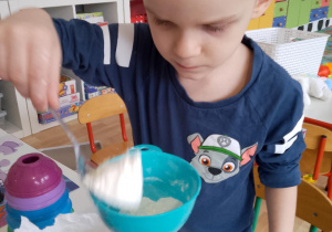 Sala przedszkolna. Chłopiec za pomocą łyżki i lejka, wsypuje mąkę do plastikowej butelki.