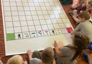 Dzieci siedzą na dywanie przed matą do kodowania, na której ułożone są kolorowe obrazki.