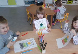 Troje dzieci przy stolikach pokazują swoje narysowane prace- zimowe czapki.