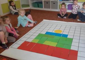 Dzieci siedzą na dywanie wokół maty do kodowania, na której dzieci utworzyły z kolorowych kwadratów zimową czapkę.